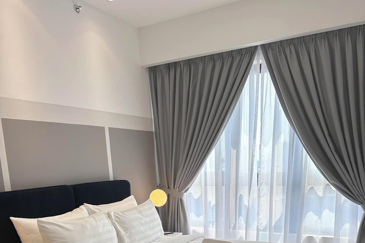 Una habitación de hotel moderna con una cama grande, una mesita de noche, una lámpara y vistas a través de cortinas translúcidas.