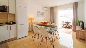 A modern kitchen with white cabinets and a built-in oven. There is a dining area with a glass table and chairs, and a small wooden side table with a plant. - Seaside Getaways by Interhome (Armação de Pêra)