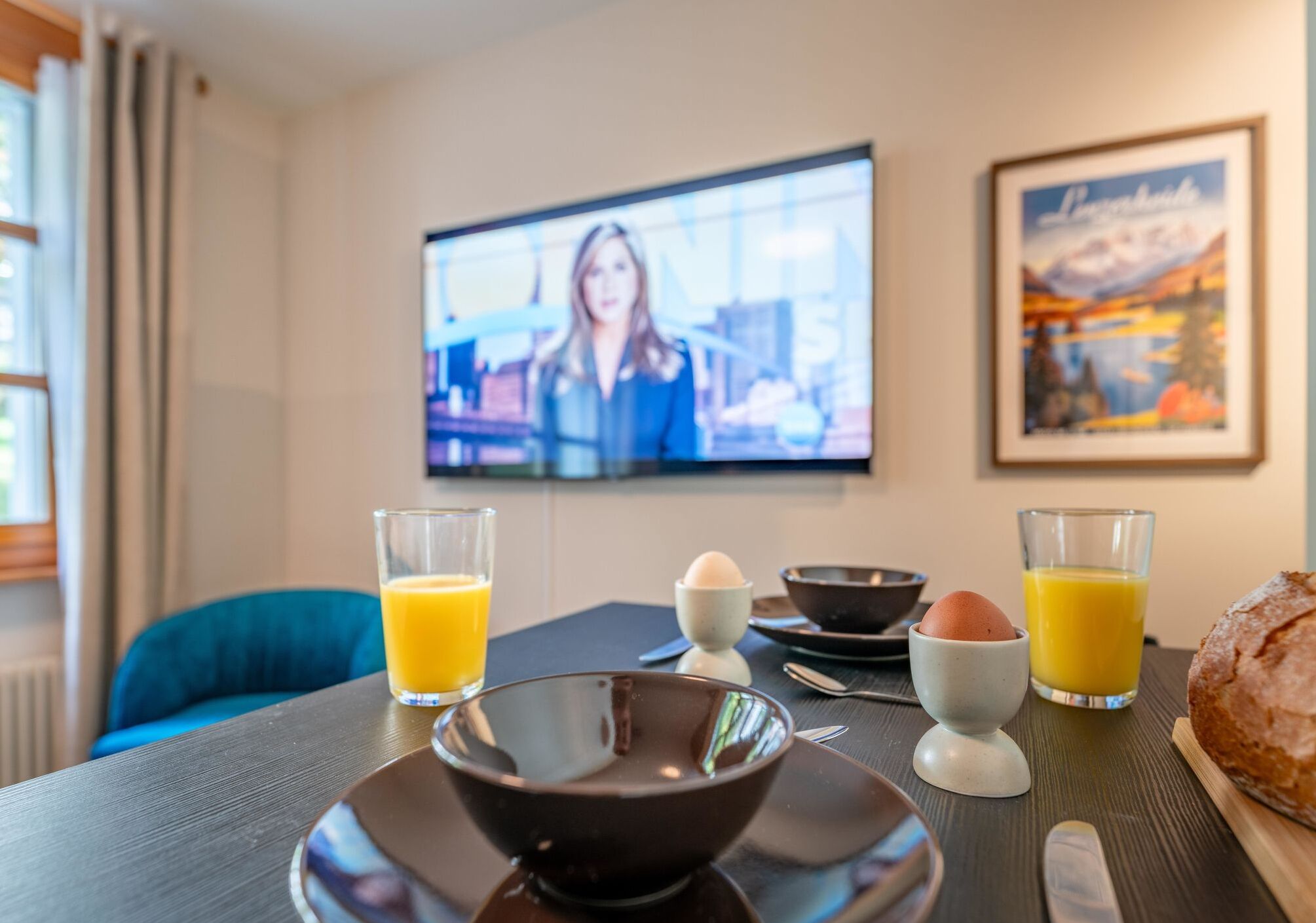 Una preparación para el desayuno con un televisor mostrando un noticiero, un cuadro en la pared y una hogaza de pan sobre la mesa.
