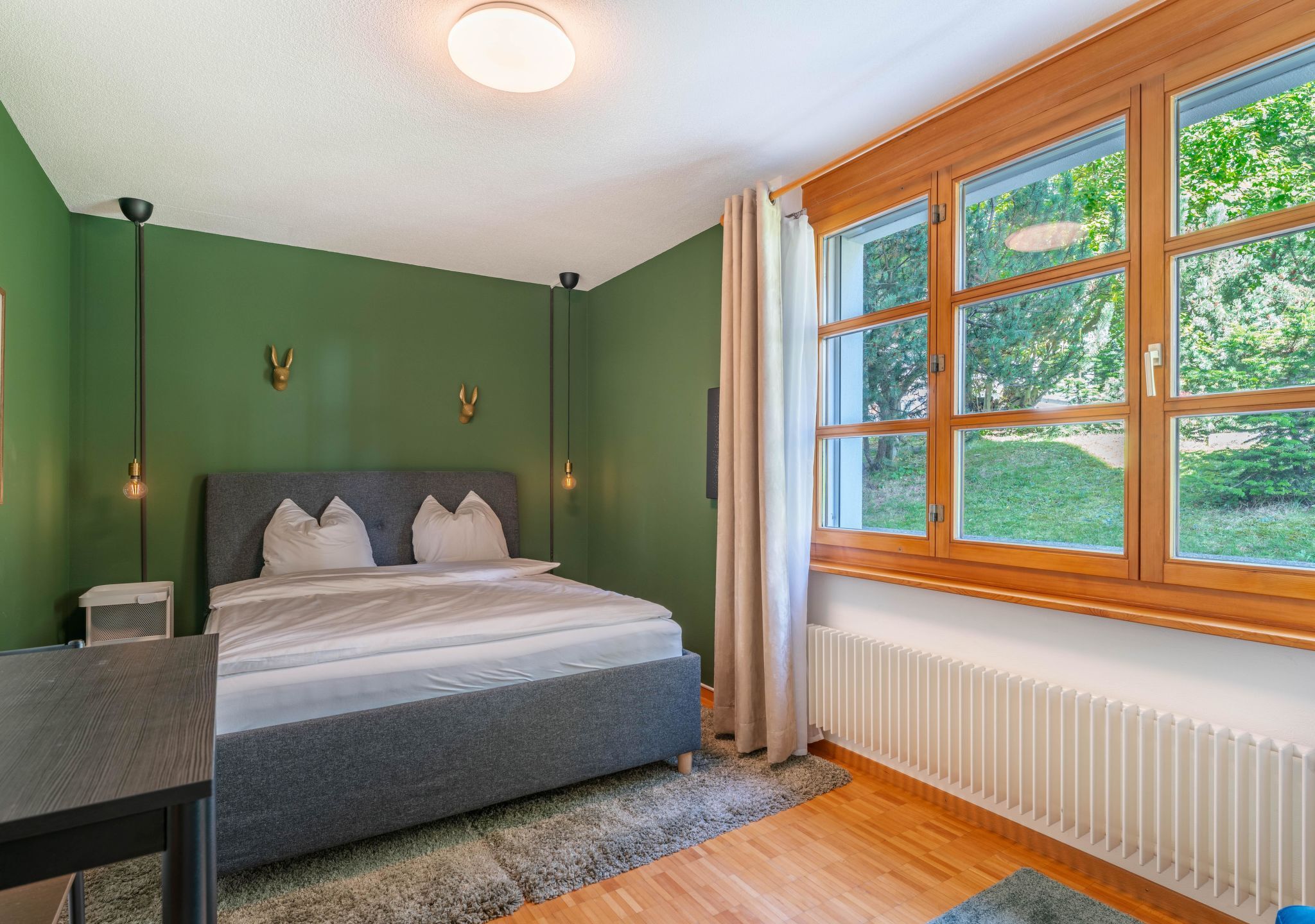Quarto com cama, mesa de cabeceira, luminária e janela com vista para vegetação.