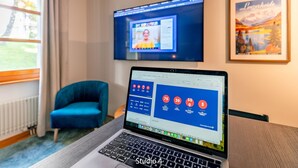 A MacBook Pro on a table with a spreadsheet open, a TV displaying a video call, a blue chair, and a framed picture on the wall. - Steil Alva Studios Smit by Interhome (Lenzerheide)