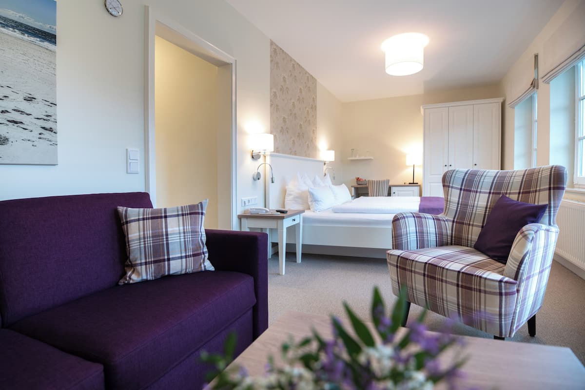 Ein Hotelzimmer mit einem Bett, einem violetten Sofa, einem Karosessel, einem kleinen Tisch und einer Wandlampe.