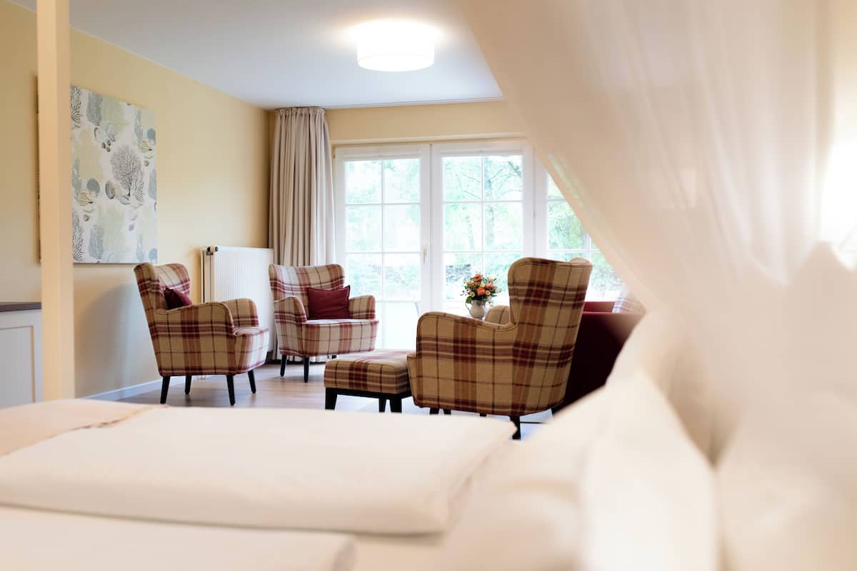 Ein Zimmer mit einem Bett, zwei karierten Sesseln, einem Heizkörper und einem Fenster mit Vorhängen.