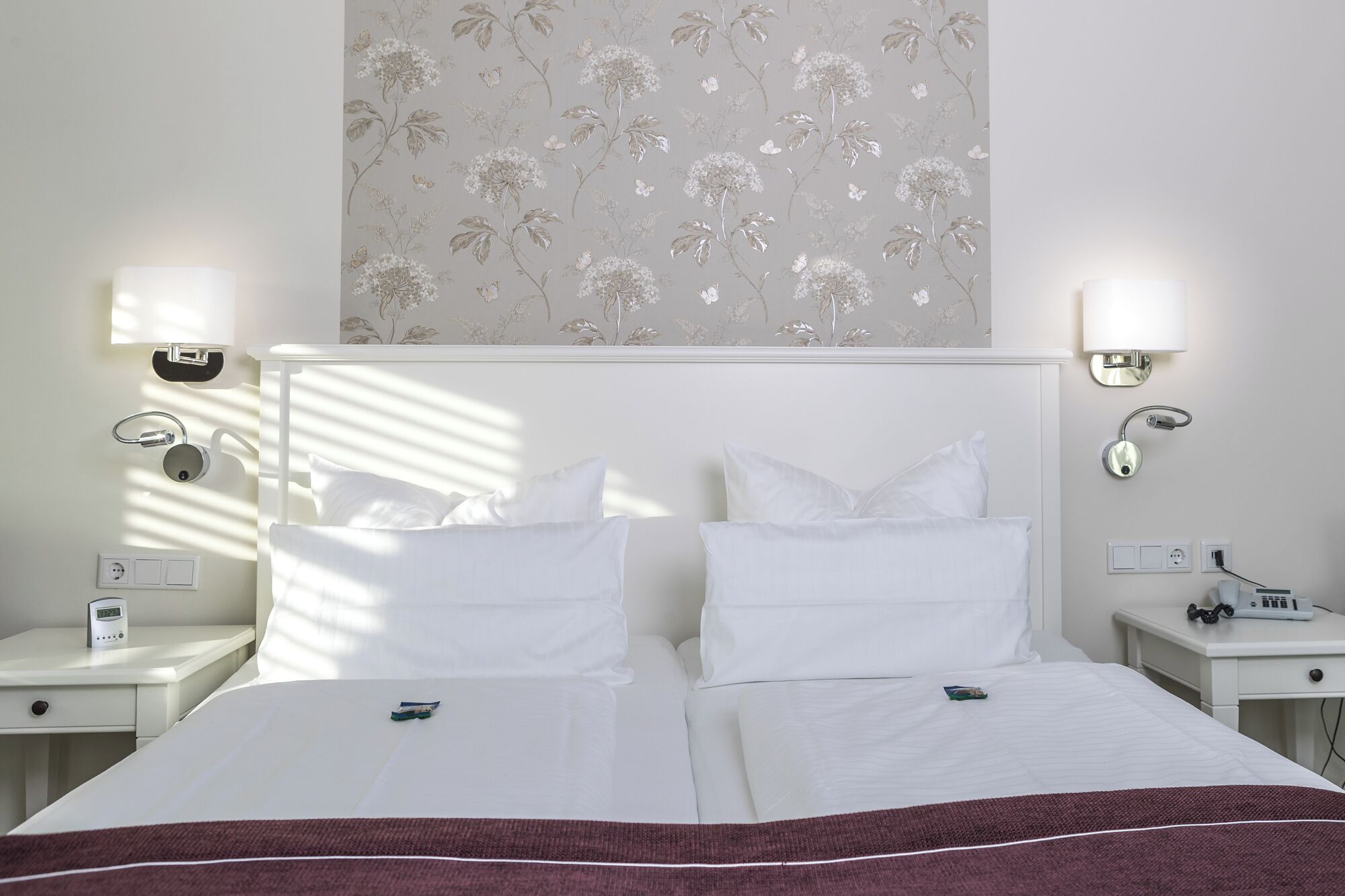 A hotel room with two beds, a nightstand with a lamp, a wall-mounted outlet, and a power strip.