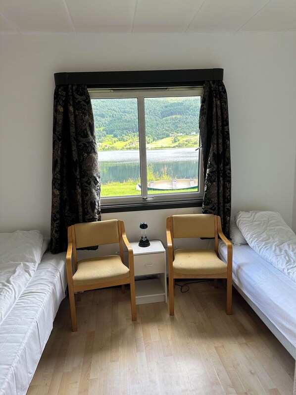 A room with two single beds, a small desk with a lamp, two chairs, and a window with curtains.
