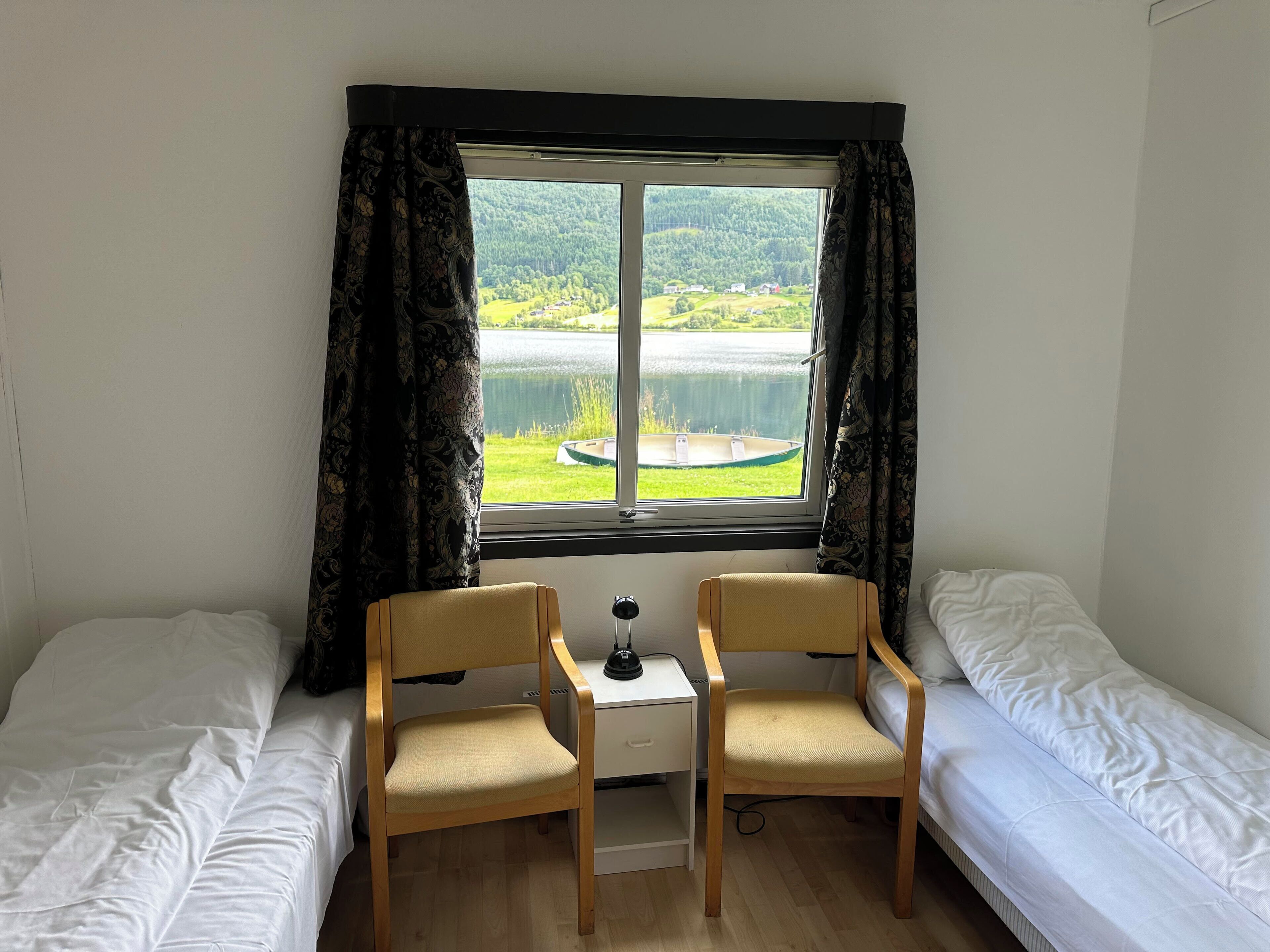 A room with two single beds, a small desk with a chair, and a window offering a view of a lake and greenery.