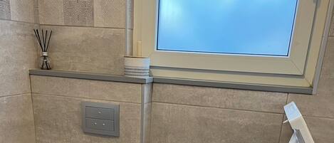 Un bagno con il water, un cestino, una finestra e una lavatrice.