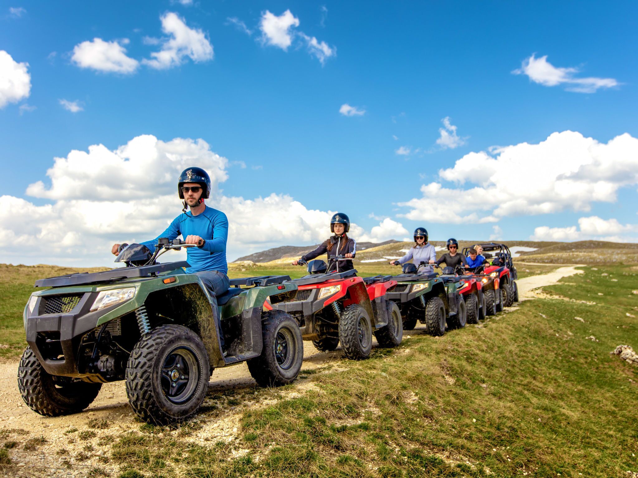 A group of people riding ATVs on a dirt path with grassy fields and hills in the background.