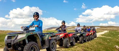 A group of people riding ATVs on a dirt path with grassy fields and hills in the background.