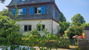Une maison à deux étages, dotée d’un toit à forte pente, de nombreuses fenêtres avec volets et d’une cheminée bien visible.