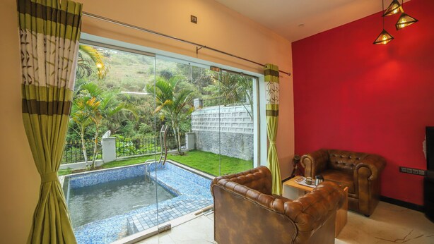 A living room with a red wall, a brown leather sofa, a glass-top coffee table, and a view of a swimming pool and garden. - Rain Rock Villa (Yercaud)