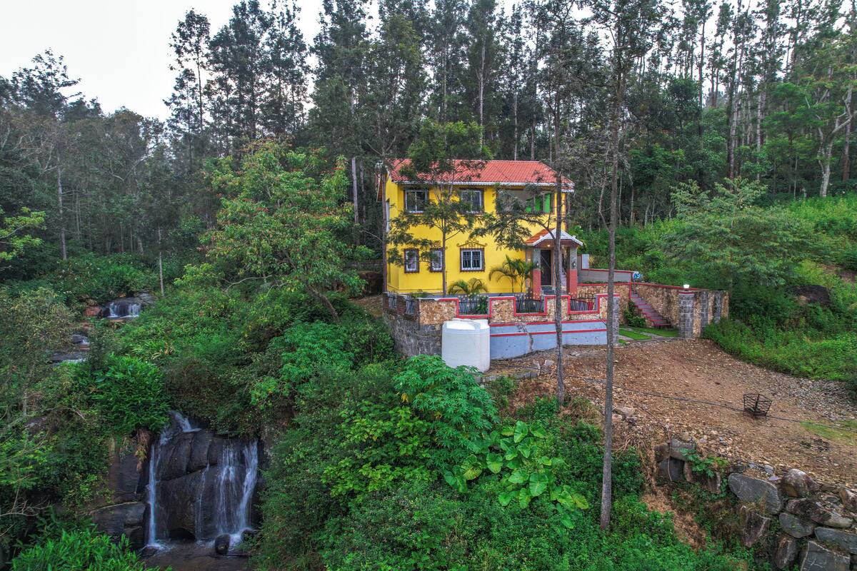 A yellow house with a red roof surrounded by lush greenery and a waterfall.