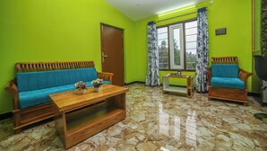 Una habitación con un sofá de madera, una mesa de centro de madera, un sillón de madera, una mesita con un florero y una ventana con cortinas.
