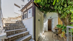 A narrow outdoor corridor with stone steps leading to a door, a small patio area with a table and chairs, and a tree providing shade. - Casa Lorenzo OG - Ap 2 - 1.kat (Premantura)