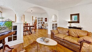 A spacious living room with a leather sofa, wooden coffee table, and a dining area with a chandelier. - Historic Spanish Charm + Scenic View | Cozy Casita (Birmingham)