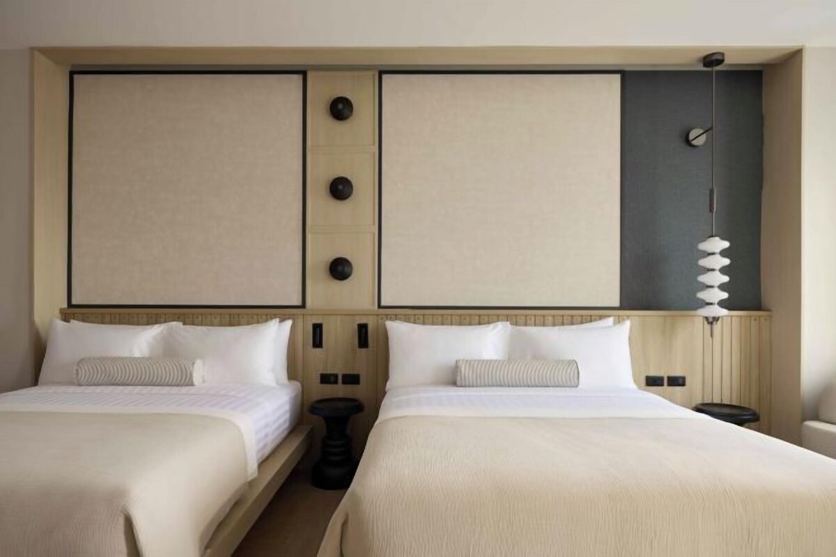 A hotel room with two beds, a large wall panel with buttons, a hanging light fixture, and a small round table.