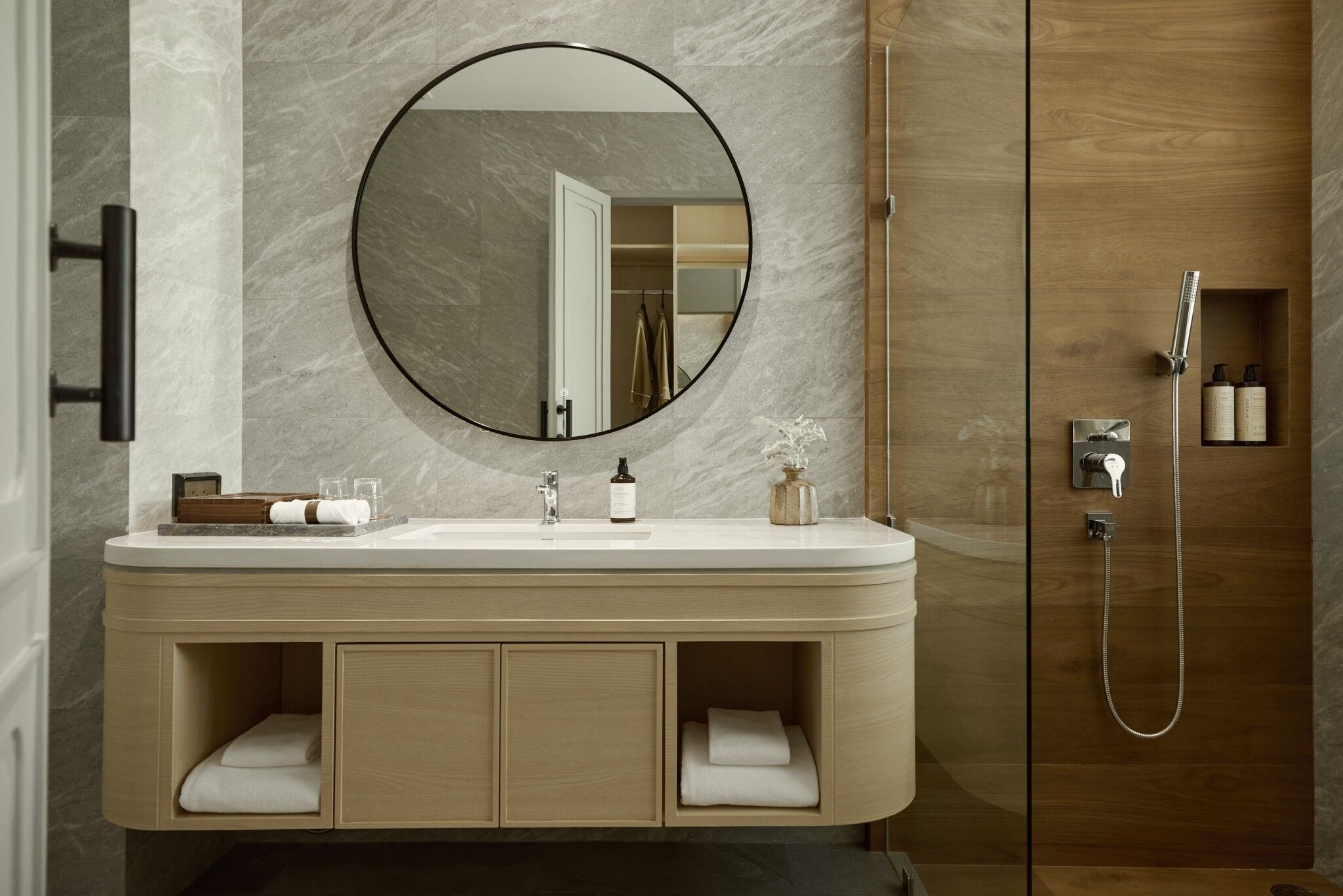 A modern bathroom with a marble wall, a round mirror, a sink with toiletries, and a shower area.