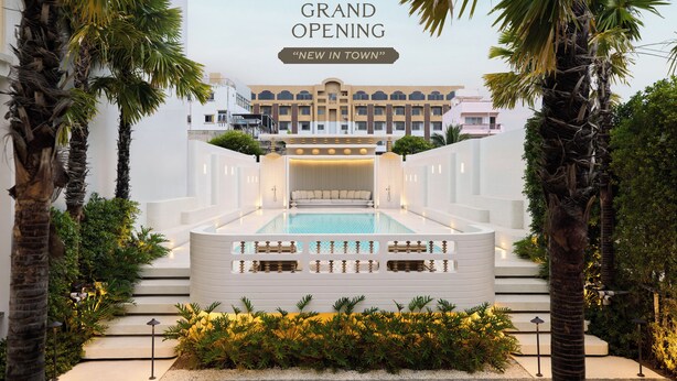 Een modern hotel met een zwembad, palmbomen en een bord met 'Groot Openingsfeest 2024'.