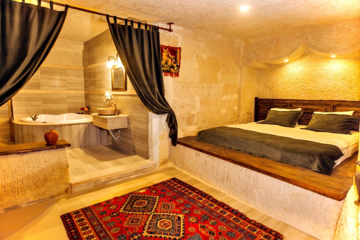 A bedroom with a wooden headboard, a large bed with pillows, a nightstand, a mirror, a sink, and a shower area.