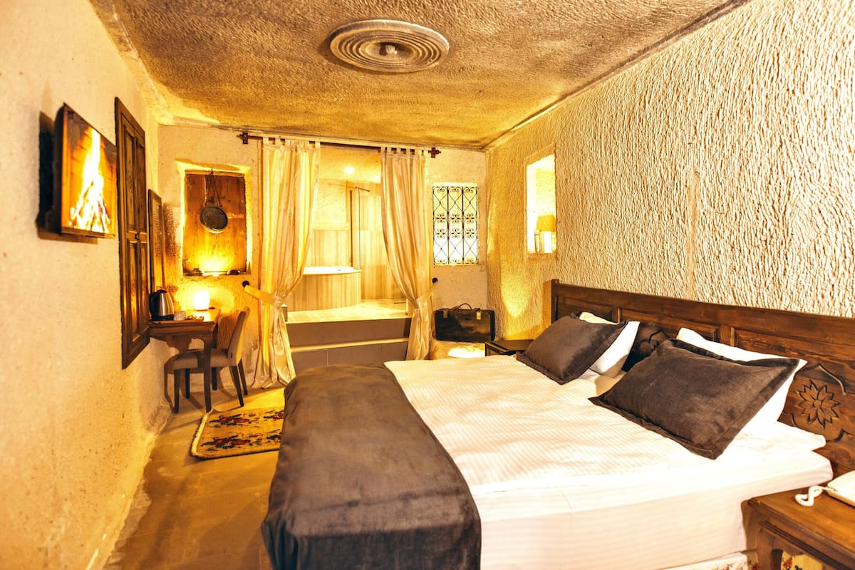 A bedroom with a large bed, a desk with a chair, a small table with a lamp, and a window with curtains.