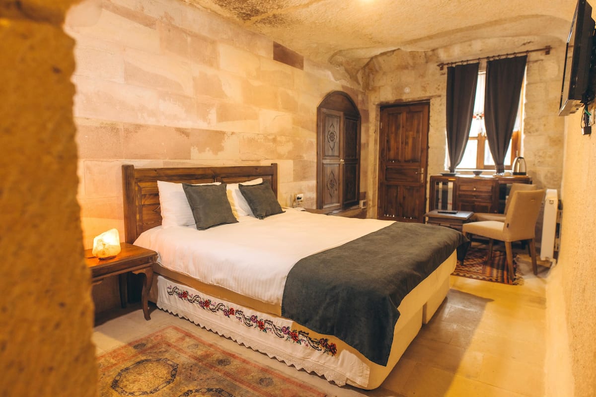A bedroom with a large bed, wooden headboard, a nightstand with a lamp, a desk with a chair, and a wardrobe.