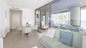 A modern living room with a green sofa, a dining area with white chairs, and a large glass wall offering a view of the building's interior. - Apartamento Infinium 3 - Costa CarpeDiem (Calpe)