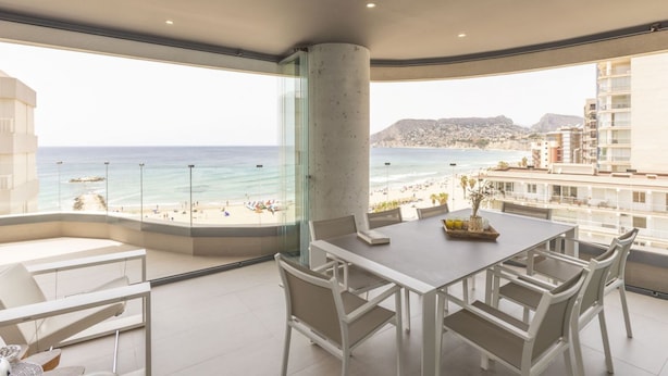A modern dining area with a large table and chairs, overlooking a beach and cityscape. - Apartamento Infinium 3 - Costa CarpeDiem (Calpe)