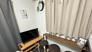 Un petit salon avec un téléviseur à écran plat, un banc en bois, deux tabourets, un ventilateur blanc et une horloge au mur.