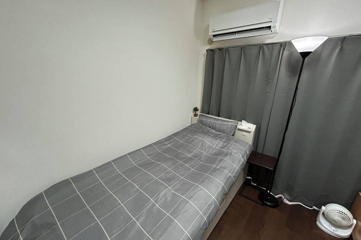 A small, single-bed room with a grey checkered bedspread, a bedside table with a fan, and an air conditioning unit mounted on the wall.