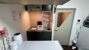 Une cuisine compacte équipée d’un évier, d’un four à micro-ondes et d’un petit plan de travail. On y trouve un vase blanc rempli de fleurs sur le plan de travail, et un haut-parleur blanc posé sur le sol.
