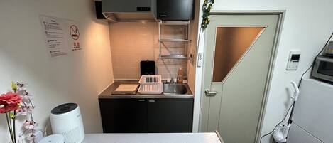 A compact kitchen with a sink, a microwave, and a small countertop. There is a white vase with flowers on the counter, and a white speaker on the floor.