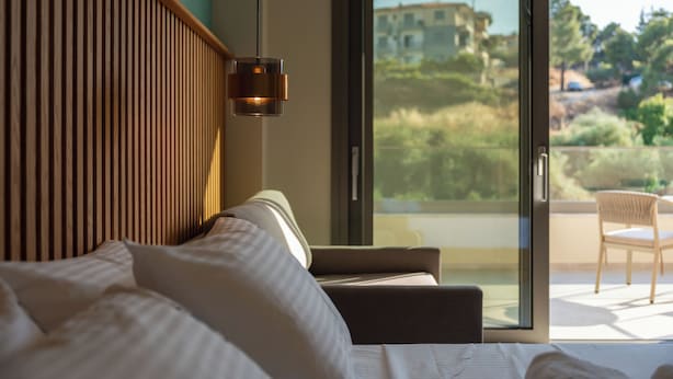 Una habitación de hotel con cama, sofá, lámpara de pared y puerta corrediza de vidrio que da a un espacio exterior con una silla.