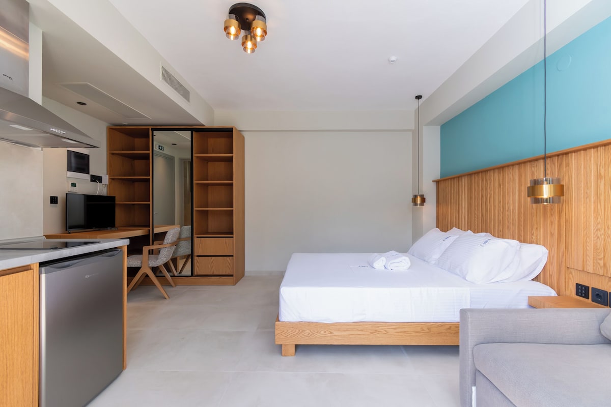 A modern hotel room with a bed, a sofa, a desk with a computer, a wooden wardrobe, and a kitchen area.