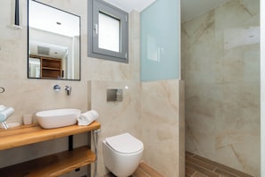 A modern bathroom with a marble wall, a wooden vanity, a white sink, a toilet, and a glass shower enclosure. - HP Family Suites (Sithonia)