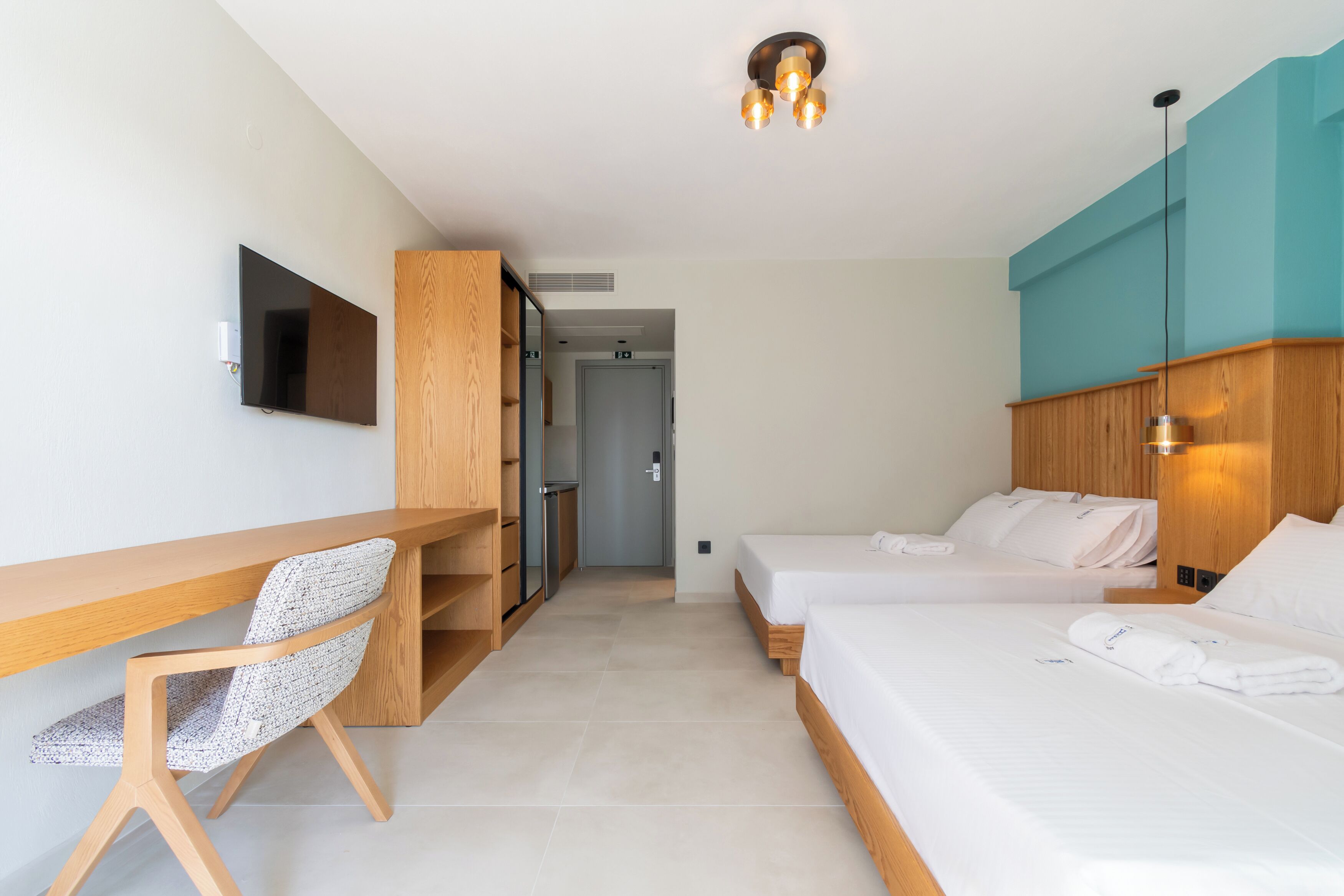 Una habitación de hotel moderna con un escritorio de madera, dos camas, un televisor y un pasillo que conduce a una puerta.