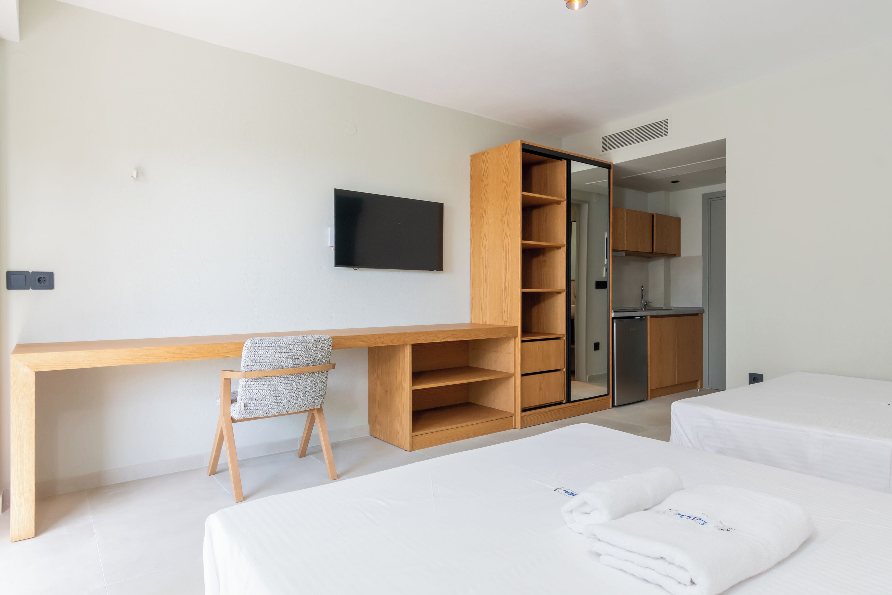 A modern hotel room with a wooden desk, a TV, a kitchenette, and two beds.