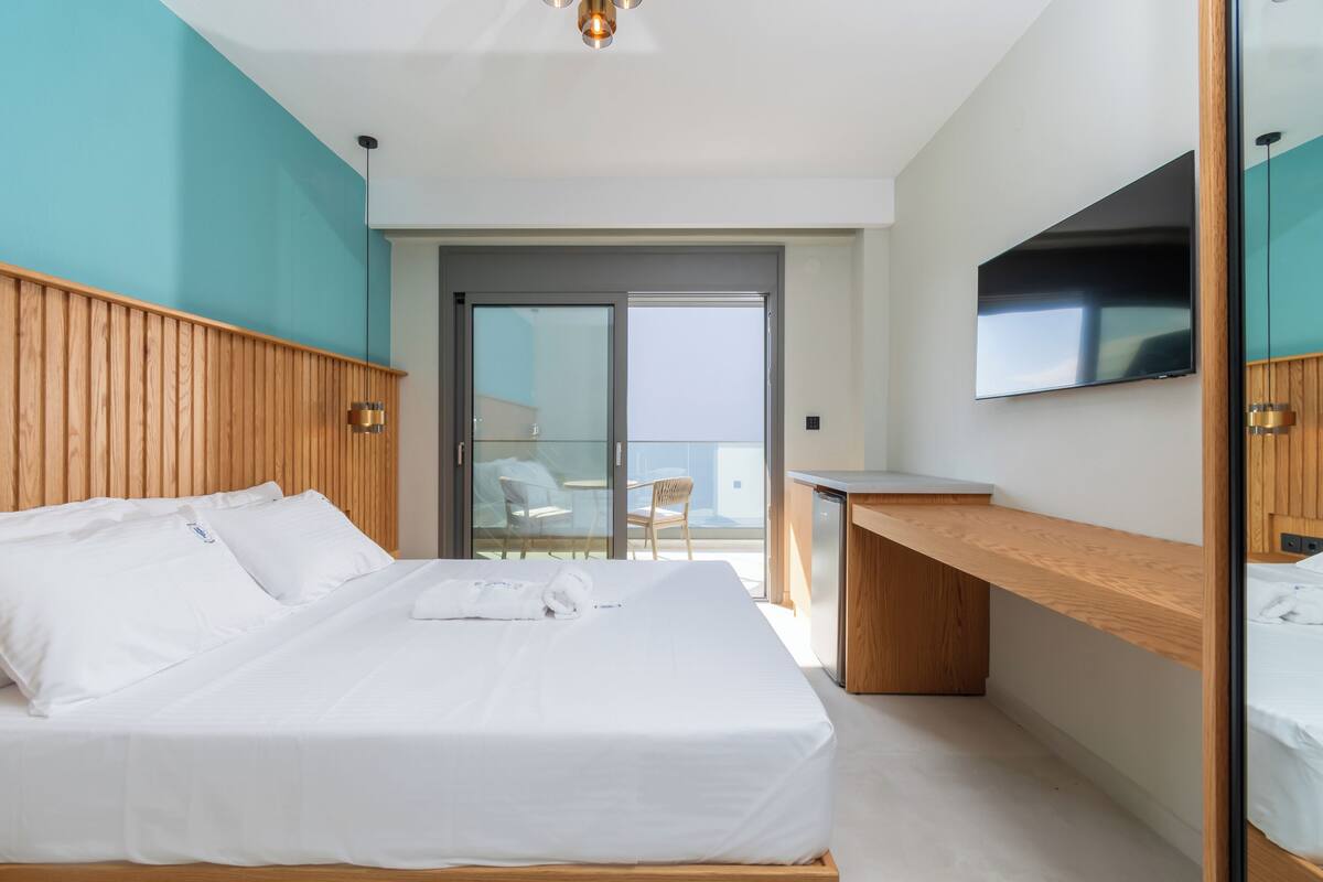 A hotel room with a large bed, a desk, a flat-screen TV, and a sliding glass door leading to a balcony with a chair.
