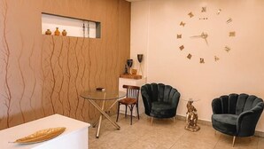 A modern room with a round glass table, two dark armchairs, a decorative wall clock, and a small shelf with vases.