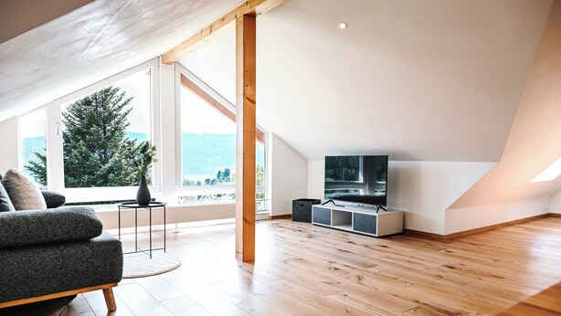 A modern loft with a large window, wooden flooring, a grey sofa, and a TV. - LakeviewOasis: Exclusive Schnepfhalde apartment (Schluchsee)