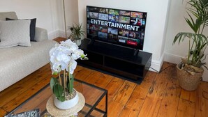 A living room with a television displaying an entertainment channel, a sofa, a coffee table with a white orchid, and two potted plants. - Amazing Victorian House, Excellent City Location (Cambridge)