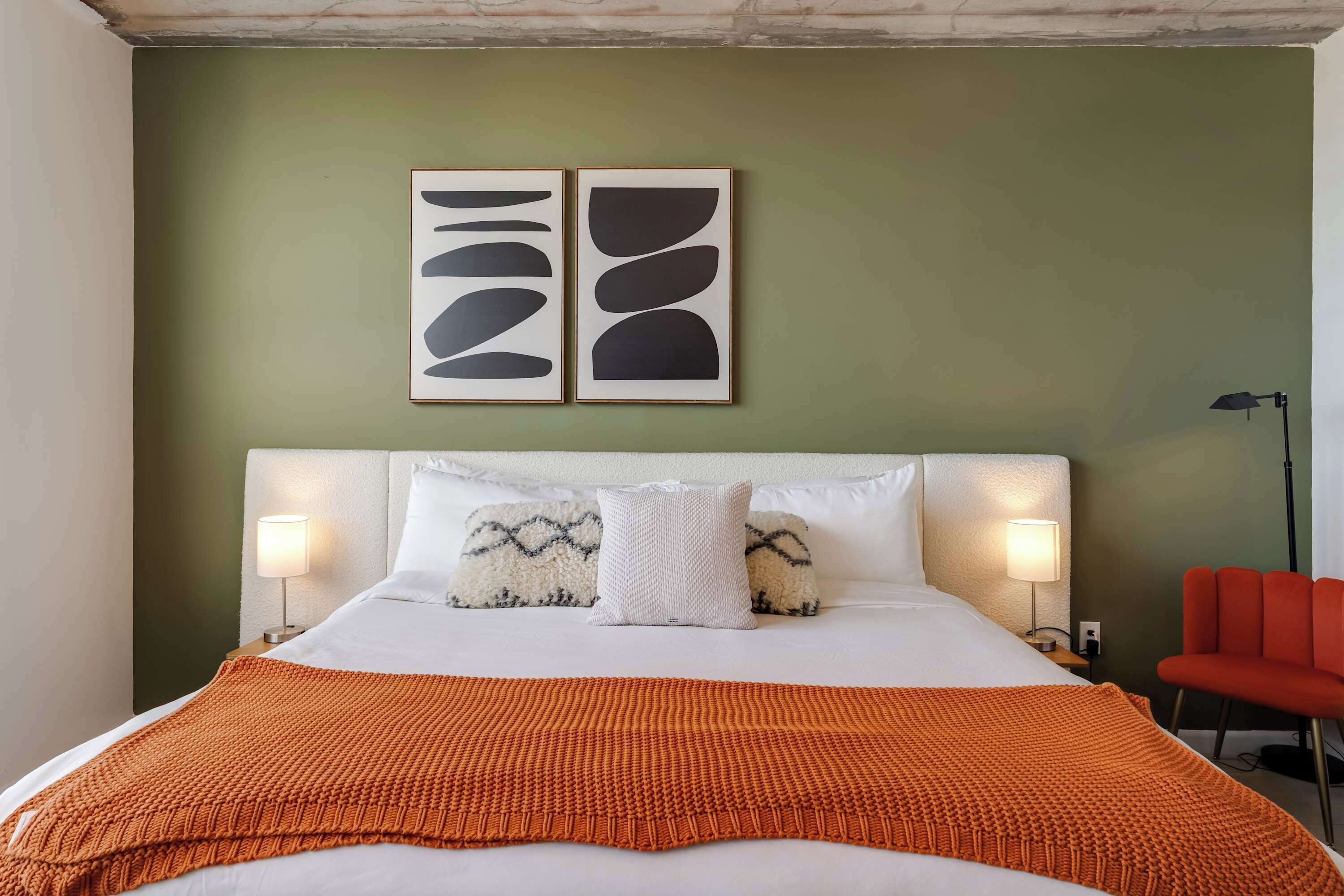 Ein Bett mit weißem Kopfteil, einem weißen Kopfkissen und einer orangefarbenen Decke. Zwei Bilderrahmen mit abstrakten Motiven sind an der Wand befestigt.