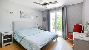 Um quarto com cama, escrivaninha, cadeira, ventoinha no teto e janela com cortinas.
