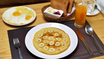 Un plato de desayuno con tortitas cubiertas de rodajas de plátano, una porción de huevos fritos, tostadas y un vaso de zumo de naranja sobre una mesa de madera.