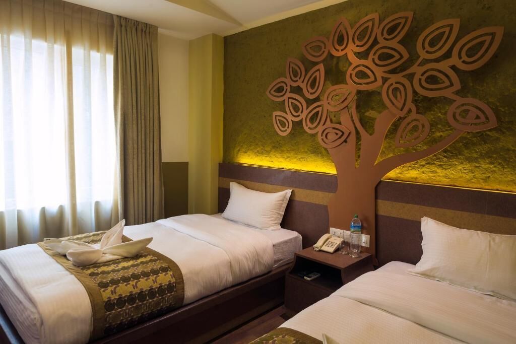 A hotel room with two beds, a decorative tree wall art, a bedside table with a phone and water bottle, and a window with curtains.