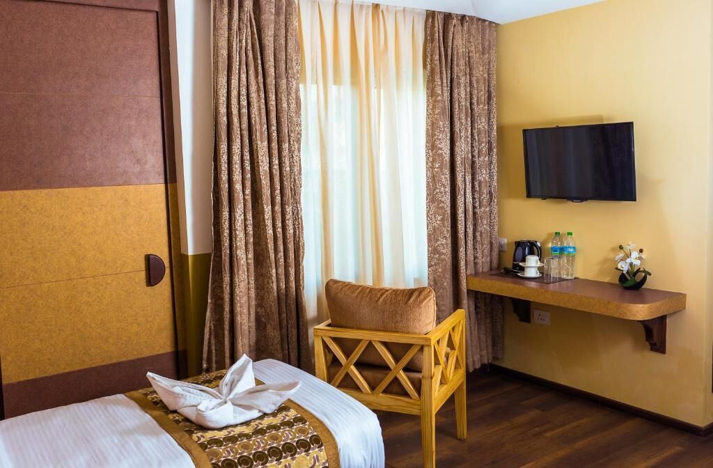 Una habitación de hotel con cama, una silla de madera, televisor, una mesita con un florero y cortinas.