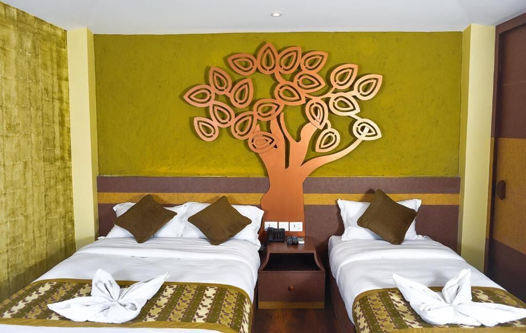 Une chambre d’hôtel avec deux lits, une décoration murale en forme d’arbre et une table de chevet en bois sur laquelle se trouvent un téléphone et des télécommandes.