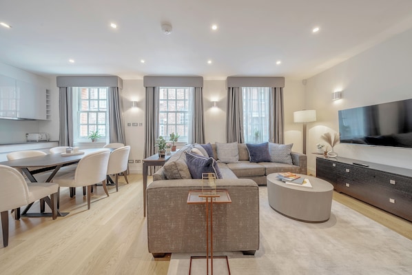 A modern living room with a sofa, coffee table, dining table, and chairs. There is a TV mounted on the wall and a lamp on a side table.