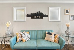 A teal sofa with geometric patterned cushions, flanked by two glass side tables with decorative vases, against a wall with framed artwork and a city skyline sign.