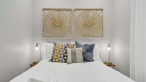 A neatly made bed with white bedding, two wall-mounted light fixtures, and two wall-mounted decorative panels.