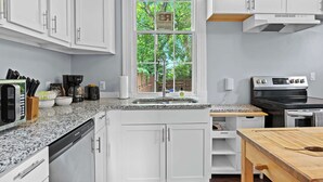 A modern kitchen with white cabinets, a granite countertop, a built-in dishwasher, a stove, and a window with a view of trees. - 3BR Relaxing Home in Louisville's Prime Location! (Louisville)